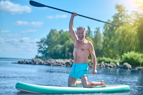 Life style. Happy confident gray-haired bearded man with raised oar in hand kneeling on board on water on sunny day