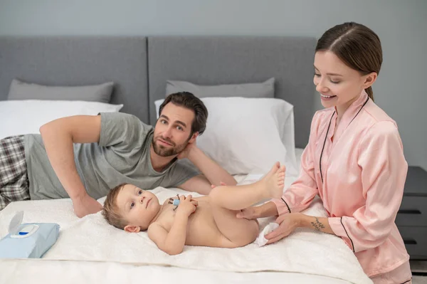 Baby skin care. Smiling young woman touching babys skin calmly lying on bed and bearded man looking at camera