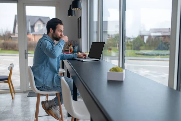 Short break. Young adult man drinking coffee sitting sideways to camera at table with laptop looking out window