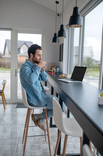 咖啡时间 一个留着胡子的年轻人侧坐着喝咖啡 带着笔记本电脑在靠近玻璃墙的房间里拍照 — 图库照片