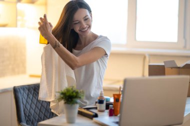 Saç bakım ürünü. Masanın önünde dizüstü bilgisayar ve kozmetik ürünleriyle saçlarına keratin sıkan mutlu genç bir kadın.