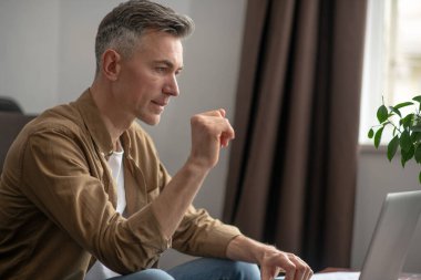 Önemli bir nokta. Kafkasyalı bir adam gündüz vakti odadaki kanepede kameraya yan oturup laptopa bakıyor.