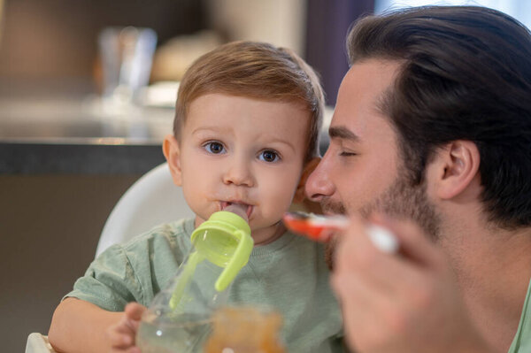 Ем. Милый маленький мальчик пьет из бутылки, пока папа кормит его.