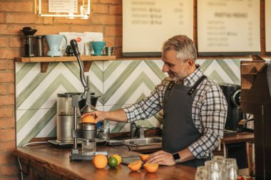 Meyve suyu. Önlüklü gülümseyen adam portakal suyu yapıyor. Mekanik meyve sıkacağı yan yana duruyor. Tezgahın arkasında kamera var.