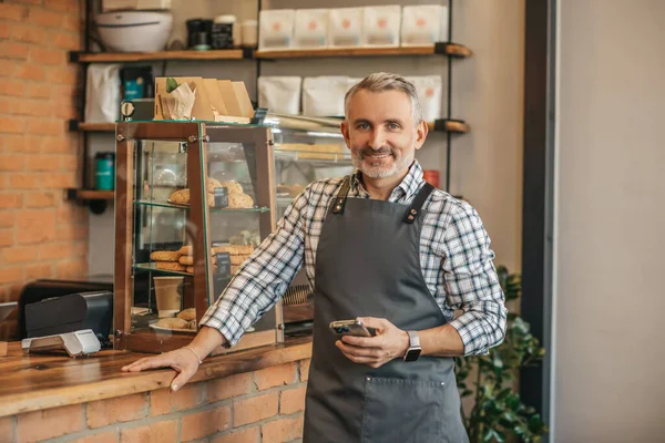 小企业 头戴围裙 自信的白发男子手握智能手机 手握酒吧柜台 站在咖啡店摄像机前微笑 — 图库照片