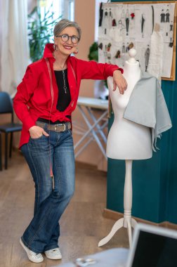 Kendine güven. Gözlüklü güzel orta yaşlı kadın atölyedeki kameraya güvenle gülümseyen mankene dokunuyor.