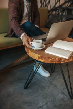 Kahve zamanı. Kanepede oturmuş laptopun yanındaki kahveye dokunan zarif bir adamın eli. Kapalı alanda kitap okuyor, yüzü yok.