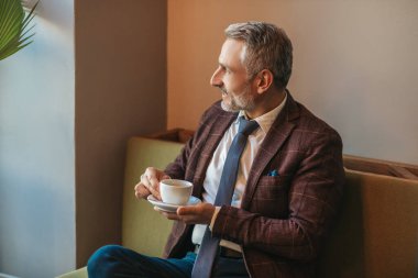 Kendine zaman ayır. Gri saçlı, zarif, orta yaşlı bir adam, elinde kahve, koltukta oturmuş, meditasyon yapıyor, içeri bakıyor.