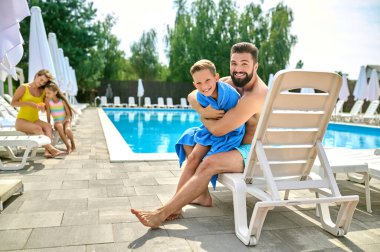 Happy young father seated in the deckchair hugging his cute son wrapped in a towel