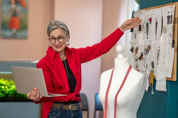 Clothes sketch. Elegant smiling woman in glasses holding laptop looking at screen pointing at clothes sketch hanging on wall
