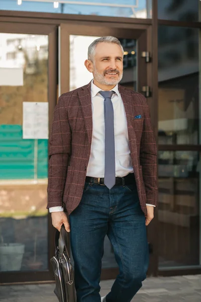 Lifestyle.Happy middle aged business man in tie with suitcase looking away walking at camera on background of office building