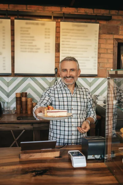 Welcome. Happy middle aged man standing at bar counter in cafe smiling holding out delicious cake on plate to camera