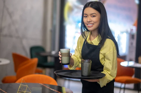 一杯卡布奇诺穿着围裙 拿着一杯卡布奇诺咖啡的漂亮的亚洲女人微笑着站在桌旁的相机前 — 图库照片