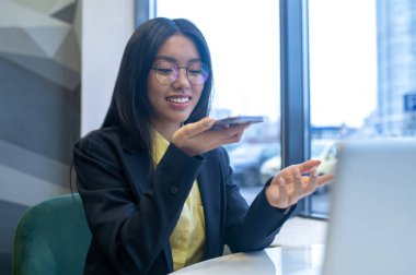 Sesli mesaj. Gülümseyen Asyalı uzun saçlı kadın çene hizasında akıllı telefon tutuyor. Gündüz penceresinin yanında dizüstü bilgisayarının başında oturuyor.