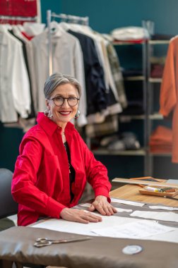 Yeni model. Gözlüklü, kendine güvenen, kameraya bakan, masada oturan, atölyedeki dokunmatik desenlere sahip bir kadın.