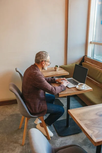 Life style. Gray-haired concentrated man in glasses typing on laptop sitting at table in cafe facing window