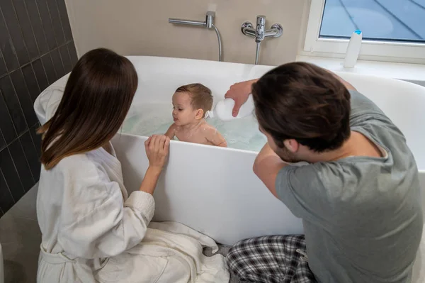 Care. Woman and man crouched sideways to camera carefully washing small cute baby sitting in foam in bath