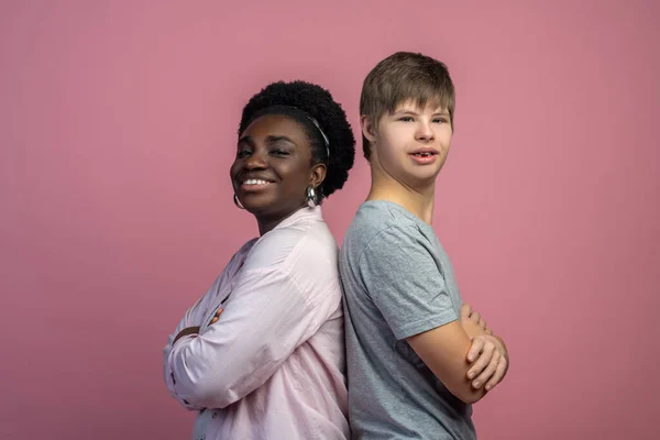 Confidence. Dark-skinned woman and caucasian guy with down syndrome standing back to back with hands folded smiling confidently at camera