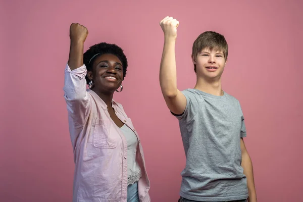 Team. African american woman and caucasian guy with down syndrome with raised fist looking at camera standing on light background