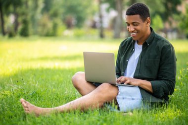 Keyfin yerinde. Güneşli bir günde çimlerin üzerinde oturup dizüstü bilgisayarda yazı yazan koyu tenli, mutlu adam.