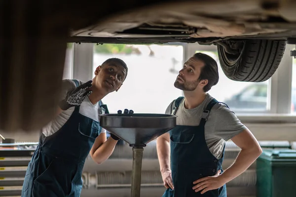 Young Auto Mechanic His Focused Colleague Examining Underside Jacked Motor — Stockfoto