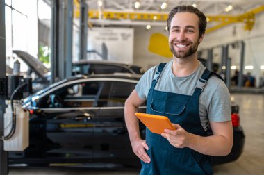 Good mood. Caucasian happy man holding tablet smiling confidently at camera standing in car service