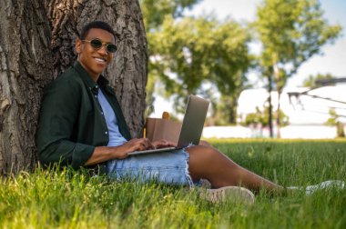 İyi günler. Güneş gözlüklü esmer tenli, kameraya gülümseyen güneşli bir günde çimlerin üzerinde çimlerin kenarında dizüstü bilgisayarla yan yana oturan.