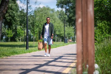 Parkta yürü. Güneş gözlüklü genç esmer adam güneşli bir günde parkta kameraya gülümsüyor.