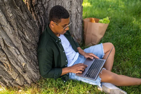 Leisure. Young man interested looking into laptop sitting on grass under tree spending time outdoors on summer day