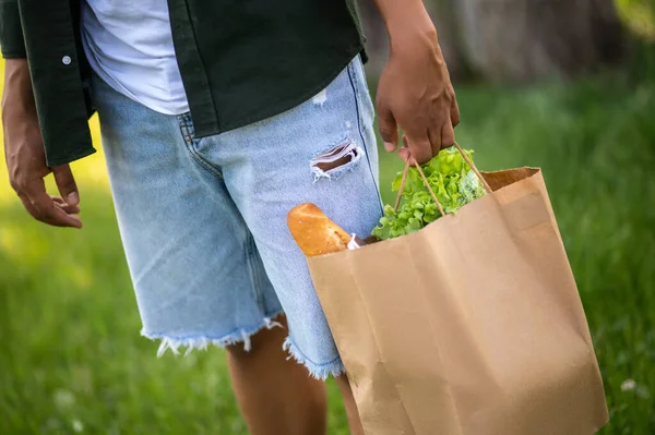 Healthy food. Male dark skinned hand holding paper bag with food outdoors on fine day without face