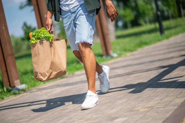Good mood. Dark-skinned man holding bag of food walking down road in park on sunny day without face