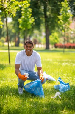 Good mood. Young dark-skinned man in protective gloves crouching picking up garbage in bag in park smiling at camera