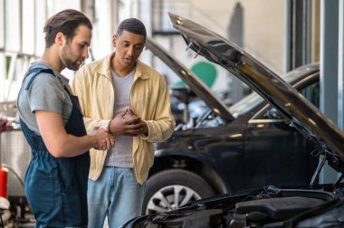 Çözüm ara. Beyaz tenli düşünceli bir tamirci kameraya yan bakıyor ve koyu tenli bir müşteri açık kaputa bakarak mantık yürütüyor.