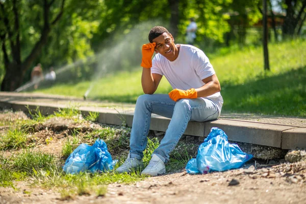 Lucky day. Smiling dark-skinned guy in protective gloves looking away sitting near packages resting in park sunny day