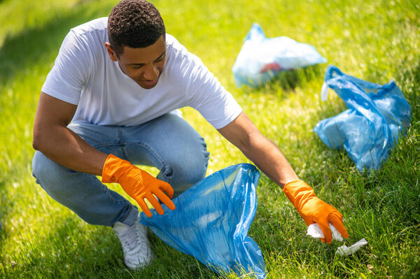 Caring, nature. Top view of dark-skinned guy in protective gloves crouched collecting garbage from grass in bag on sunny day
