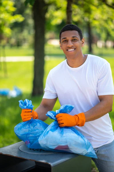 Lucky day. Dark-skinned guy in protective gloves smiling at camera holding waste bags over trash can in green park