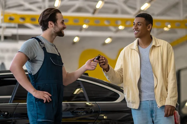 Car key. Two young adult men standing sideways to camera looking at each other touching car key at service station