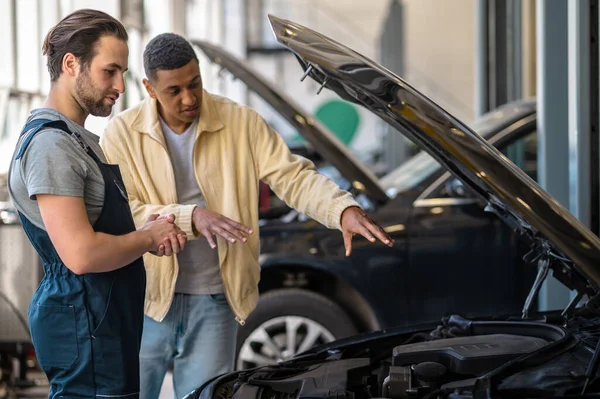 Important information. Young bearded caucasian mechanic looking attentively into car hood and dark-skinned customer telling problem standing in workshop