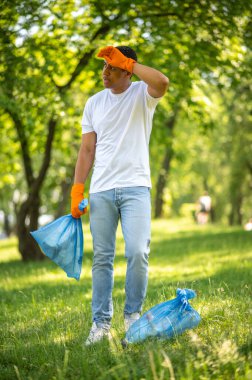 Kısa bir mola. Koyu tenli, çöp torbalı, dinlenen, yüzünü başka yöne çeviren, parkta elleriyle dokunan bir adam.