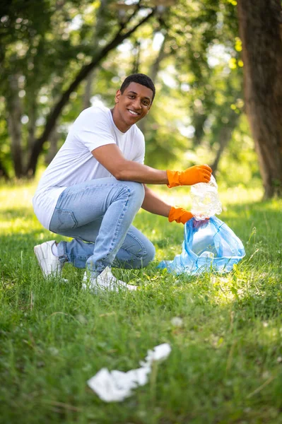 No plastic. Young dark-skinned man crouched sideways to camera picking up plastic waste smiling at camera in park