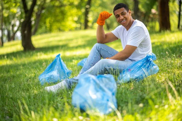 Lucky day. Smiling dark-skinned guy in protective gloves sitting near packages resting on grass in park