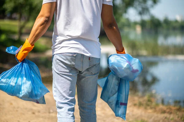 Caring for environment. Back view of man in protective gloves holding garbage bags walking near water in nature