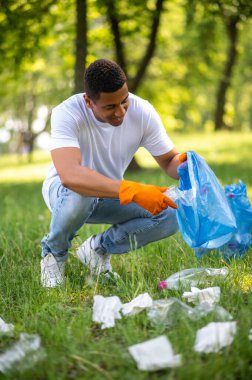 Social position. Smiling dark-skinned man in gloves crouching putting plastic in bag picking up trash in forest