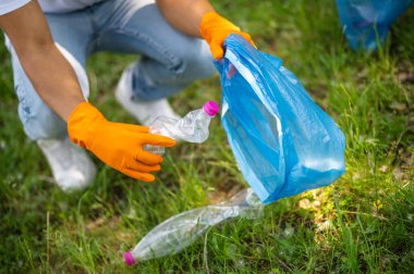 Plastic waste. Dark-skinned gloved hands holding plastic bottle near trash bag over green grass on fine day no face
