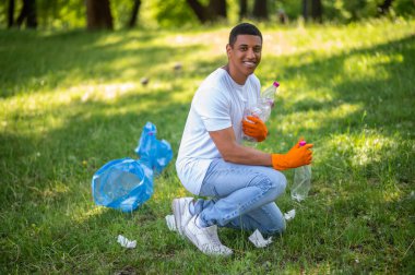 Dünya bakımı. Koyu tenli genç adam, yeşil parktaki kameraya gülümseyen plastik atıkları topluyor.