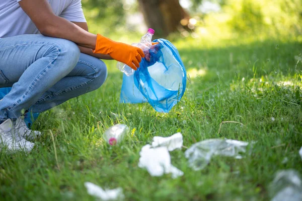 Without plastic. Dark-skinned man in gloves crouched sideways to camera with garbage bag near plastic scattered on grass outdoors no face