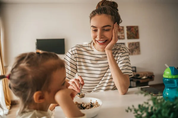 Caring Young Mother Giving Spoon Porridge Her Child Seated Kitchen — Stockfoto