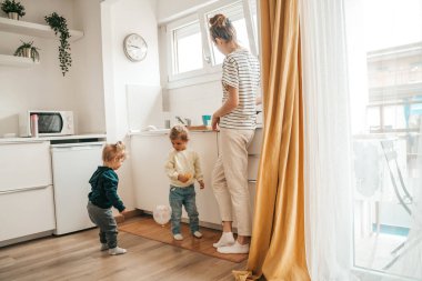 Two little children playing together with a balloon while their caring female parent cooking breakfast