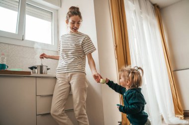 Smiling cute young woman cooking food and playing with her little child in the kitchen