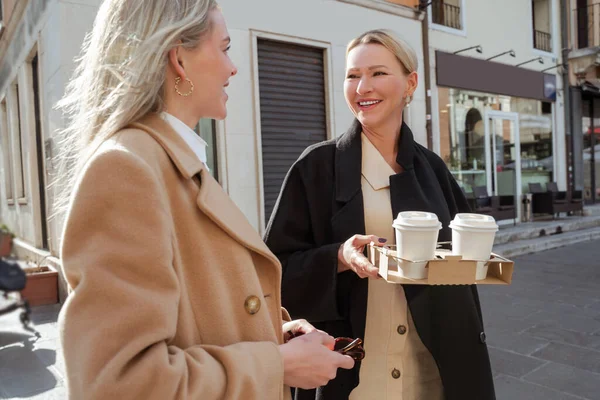 Waist-up portrait of a cheerful mature lady with takeaway coffee conversing with a blonde female